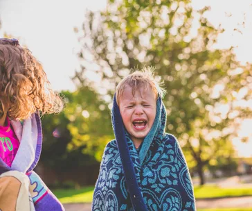A young child, wrapped in a towel, appears to be crying outdoors on a sunny day.