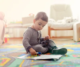 A toddler in striped pajamas sits on a colorful playmat, drawing with markers on paper in a bright room.