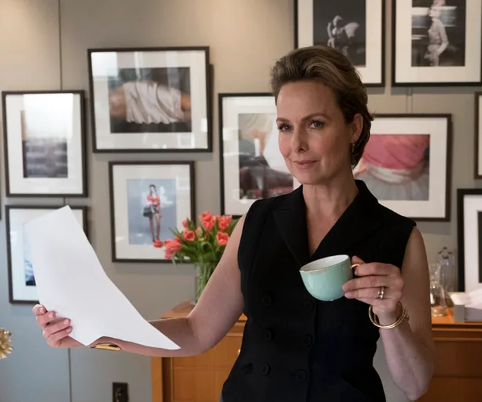 A person stands in an art-filled office, holding a document and a cup, surrounded by framed photos on the wall.