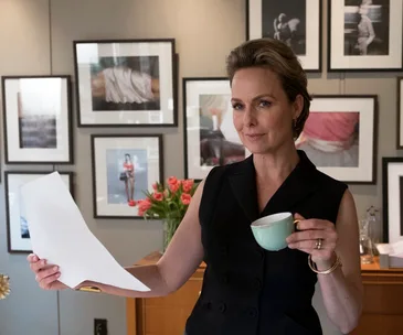A person stands in an art-filled office, holding a document and a cup, surrounded by framed photos on the wall.
