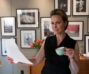 A person stands in an art-filled office, holding a document and a cup, surrounded by framed photos on the wall.