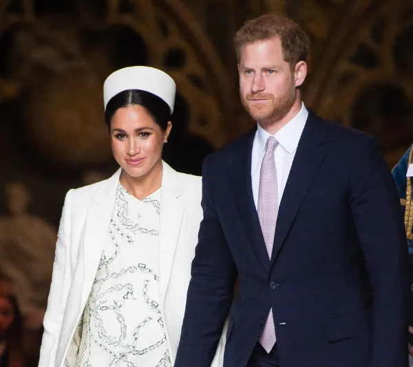 Man and woman in formal attire walking together, woman in white outfit with white hat and man in dark suit with pink tie.