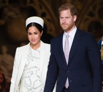 Man and woman in formal attire walking together, woman in white outfit with white hat and man in dark suit with pink tie.