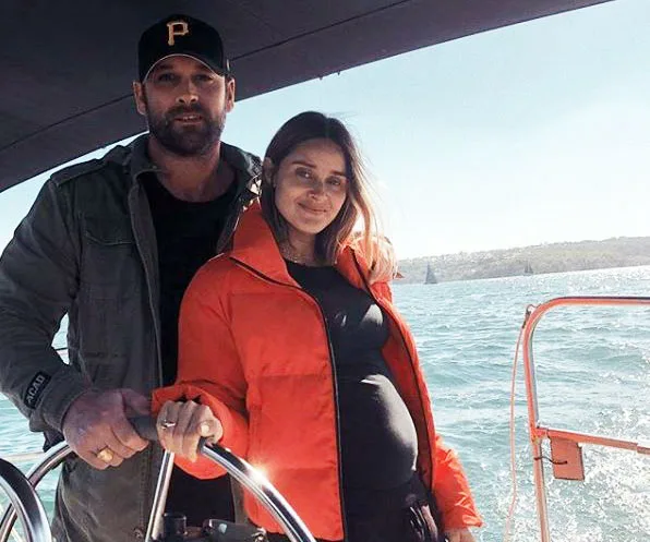 A couple on a boat; a woman in an orange jacket holds the wheel, while a man in a cap stands beside her, both smiling.