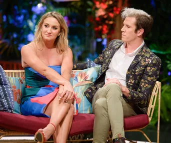 Two people sit on a colorful couch, engaged in conversation, in a vibrant, tropical setting.