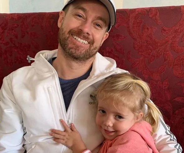 Man in white jacket and cap smiling, sitting with a young blonde girl in pink, hugging him on a red patterned chair.
