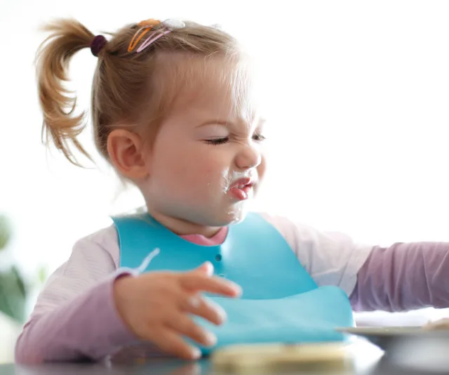 A toddler in a blue bib making a playful, scrunched face while eating at a table.