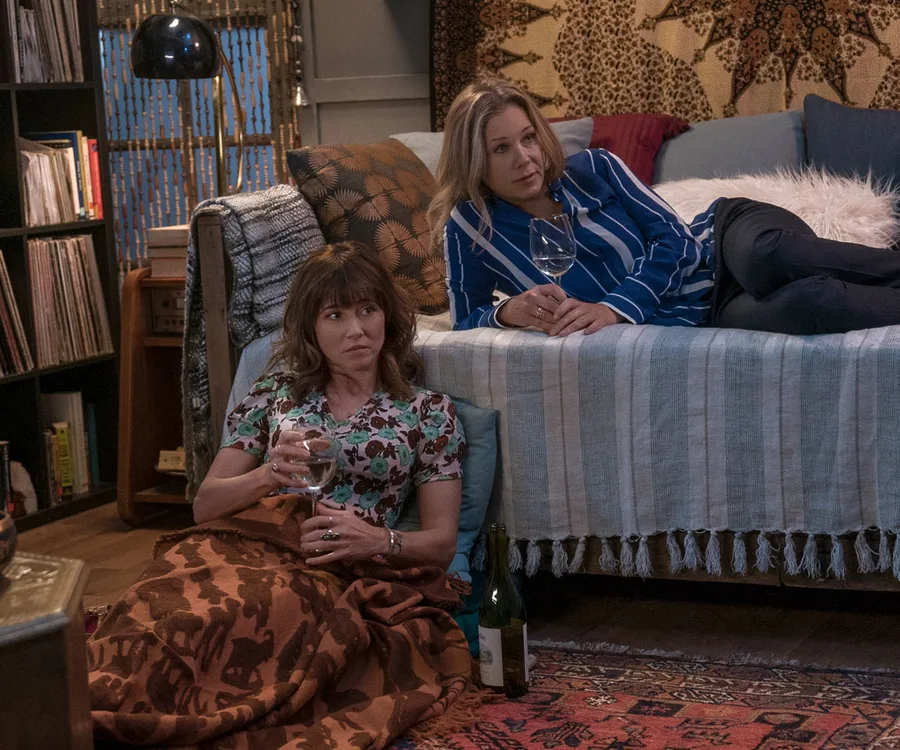 Two women sit in a cozy room, holding wine glasses. One is on a couch, the other wrapped in a blanket on the floor.