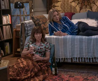 Two women sit in a cozy room, holding wine glasses. One is on a couch, the other wrapped in a blanket on the floor.