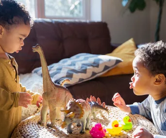 Two toddlers playing with dinosaur toys and rubber ducks on a wicker table in a cozy, sunlit living room.
