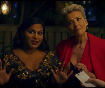 Two women talking, one with dark hair in a floral dress and the other with short hair in a red outfit, holding a phone.