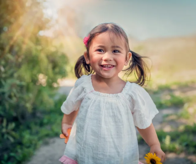 A joyful young girl with pigtails holding flowers, standing outdoors in sunlight wearing a white dress and pink hair clip.