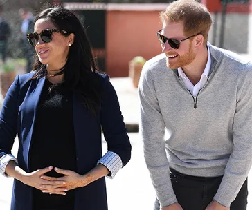 Couple smiling outdoors, woman touches belly, wearing sunglasses and navy coat; man in gray sweater, leaning slightly.