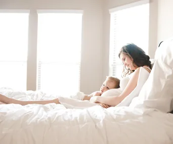 Pregnant woman and toddler cuddling on a bed, with soft daylight from the windows.