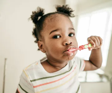 A toddler brushes their teeth with a colorful toothbrush, wearing a striped shirt, standing in a bright bathroom.