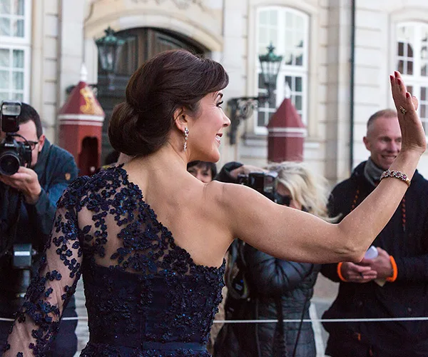 Crown Princess Mary waves to a crowd in a navy lace dress, surrounded by photographers outside a grand building.