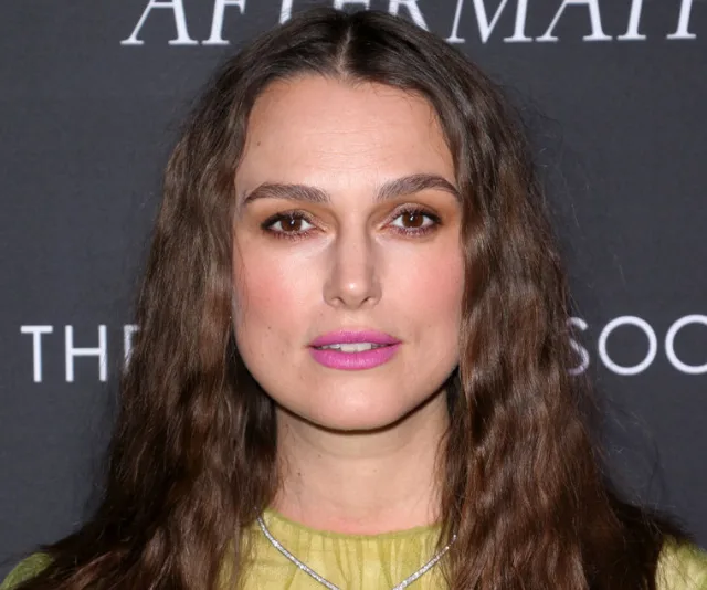 A person with long hair and bold pink lipstick poses against a dark backdrop at a formal event.