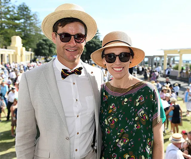 A couple in summer hats and sunglasses smiling at an outdoor event with a crowd in the background.