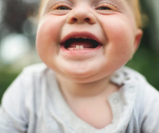 Smiling baby showing first teeth, wearing a grey shirt.