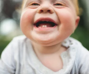 Smiling baby showing first teeth, wearing a grey shirt.