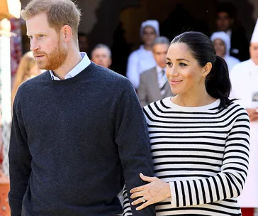 Couple holding hands, woman in striped top with henna, man in dark sweater, standing outdoors with people in background.