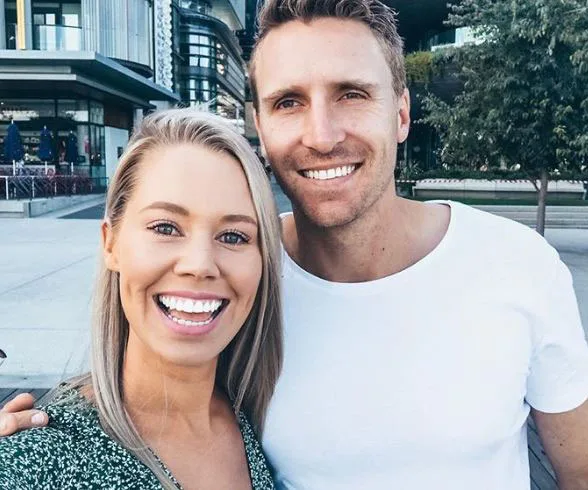 A smiling couple taking a selfie outside, with modern buildings and trees in the background.