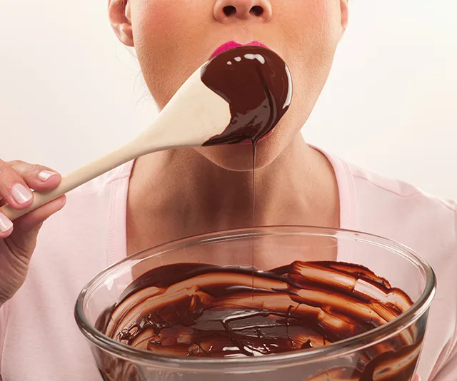Woman tasting melted chocolate with a wooden spoon from a glass bowl.