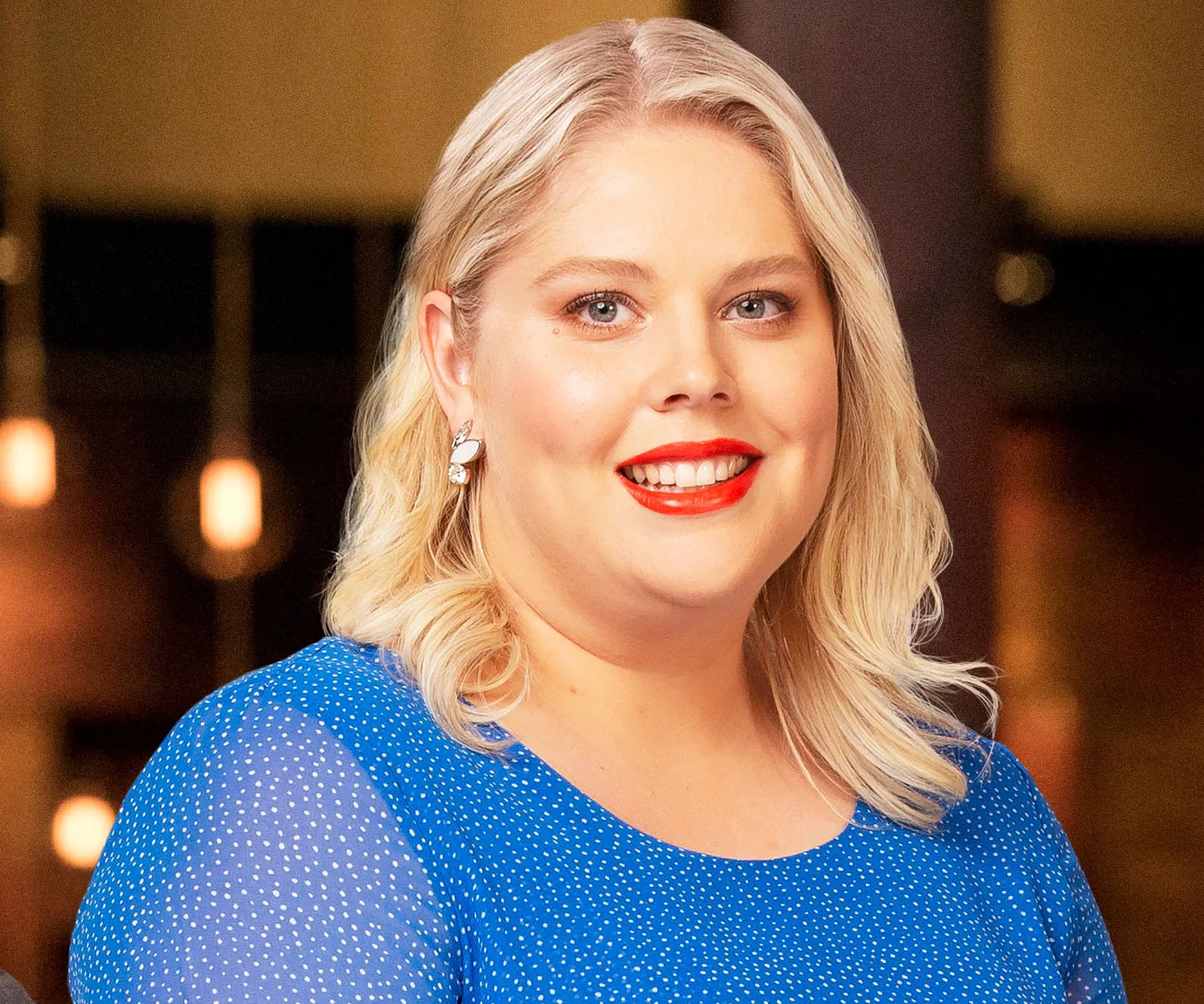 A smiling contestant from MasterChef Australia with blonde hair and a blue dress, standing indoors.