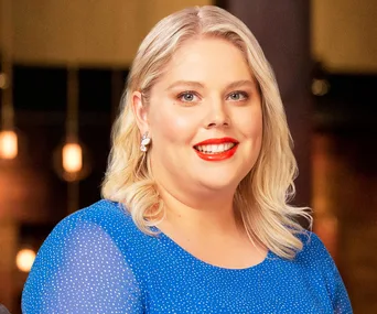 A smiling contestant from MasterChef Australia with blonde hair and a blue dress, standing indoors.