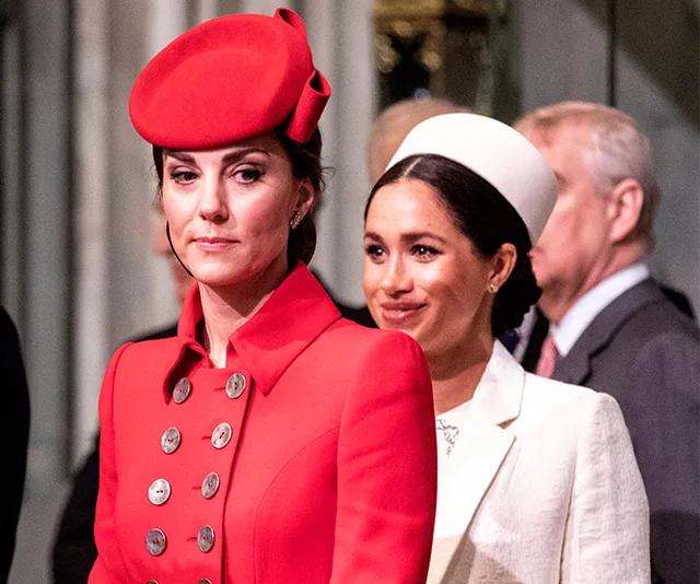 Two women at an event; one in a red coat and hat, the other in a white outfit and headpiece, with others in the background.