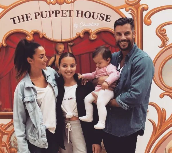 Family smiling in front of "The Puppet House" backdrop, with two adults and two children, one of whom is being held.