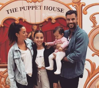 Family smiling in front of "The Puppet House" backdrop, with two adults and two children, one of whom is being held.