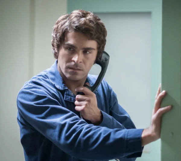 Man in blue shirt using a landline phone, appears serious, green wall in the background.