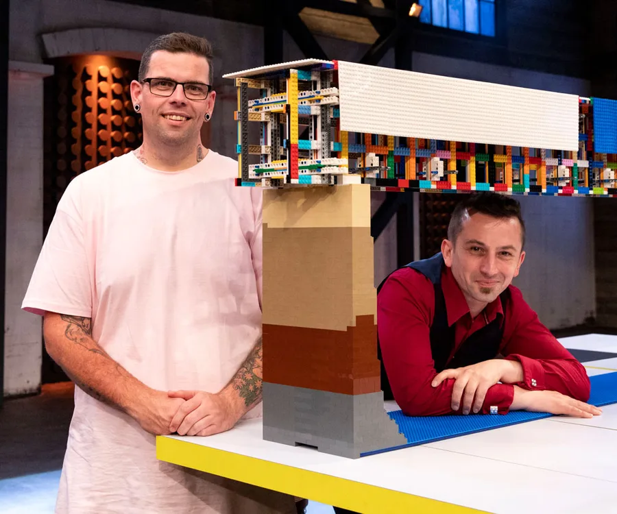 Two men with a large, colorful Lego structure on a table, one standing and one leaning on the table, smiling.