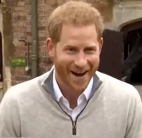 A smiling person with a beard in a light pullover, standing outdoors with a brick wall and arched doorway in the background.
