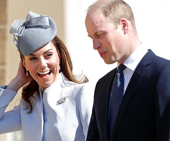 A woman in a light blue outfit and hat smiles while standing next to a man in a dark suit and tie.