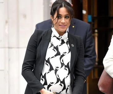 Pregnant woman in black and white floral dress and blazer, smiling outdoors, accompanied by others in the background.