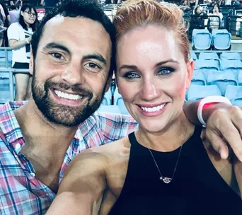 Two people smiling and posing for a selfie at a stadium event with blue seats in the background.