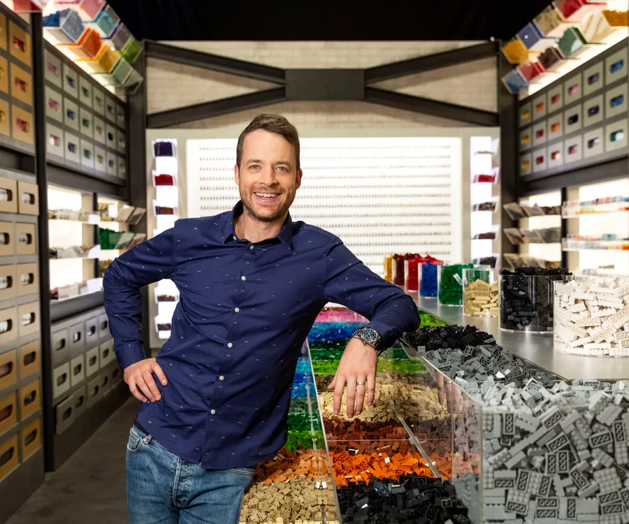 Man in a blue shirt smiles while standing in a room filled with colorful LEGO bricks on shelves and trays.