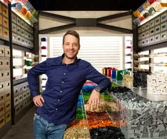 Man in a blue shirt smiles while standing in a room filled with colorful LEGO bricks on shelves and trays.