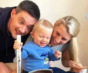 A family smiles together; a child in a blue shirt sits between two adults, enjoying a moment of joy.