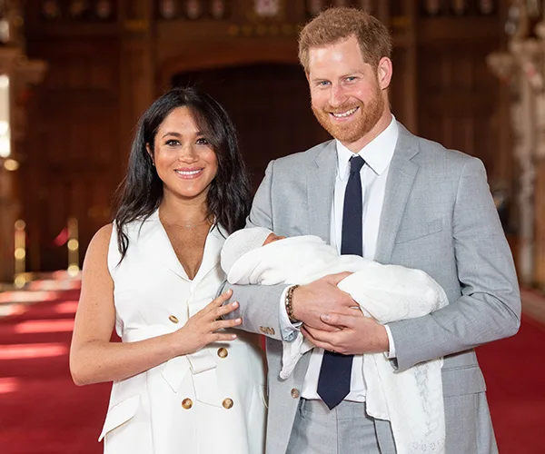 A couple smiles while holding a newborn wrapped in a white blanket, standing in an ornate hallway.