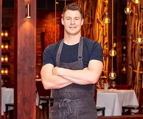Young chef in a gray apron stands confidently with crossed arms in a warmly lit restaurant setting.