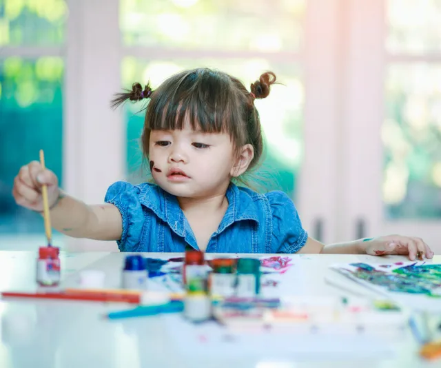 Toddler painting at a table with colorful paints, wearing a blue denim shirt, and focused on her artwork.