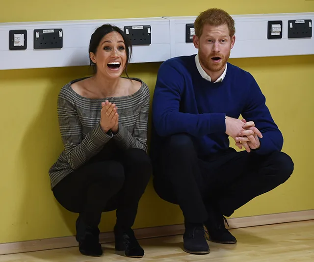 A woman and a man, both kneeling and smiling, against a yellow wall with electrical sockets.