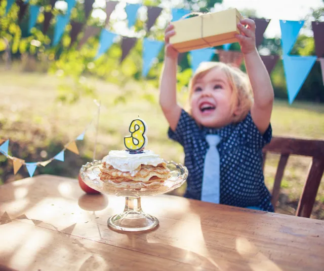 A joyful toddler celebrates a third birthday outdoors, holding a gift with a pancake cake in front.