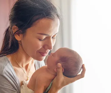 A mother tenderly holds her newborn baby close, basking in soft, natural light by a window.