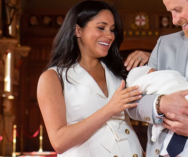 Meghan Markle smiling at her baby, who is swaddled in a blanket and held by another person, in a formal setting.