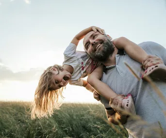 A father joyfully carries his toddler daughter on his shoulders in a sunny field.