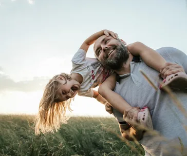A father joyfully carries his toddler daughter on his shoulders in a sunny field.